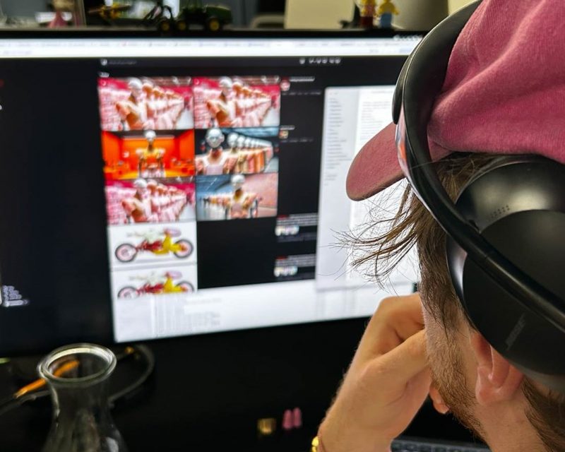 KI im Design. Eine Person mit rosa Kappe und Kopfhörern sitzt vor einem großen Monitor. Auf dem Bildschirm sind digitale Kunstwerke zu sehen. (Credits: Designworks München)